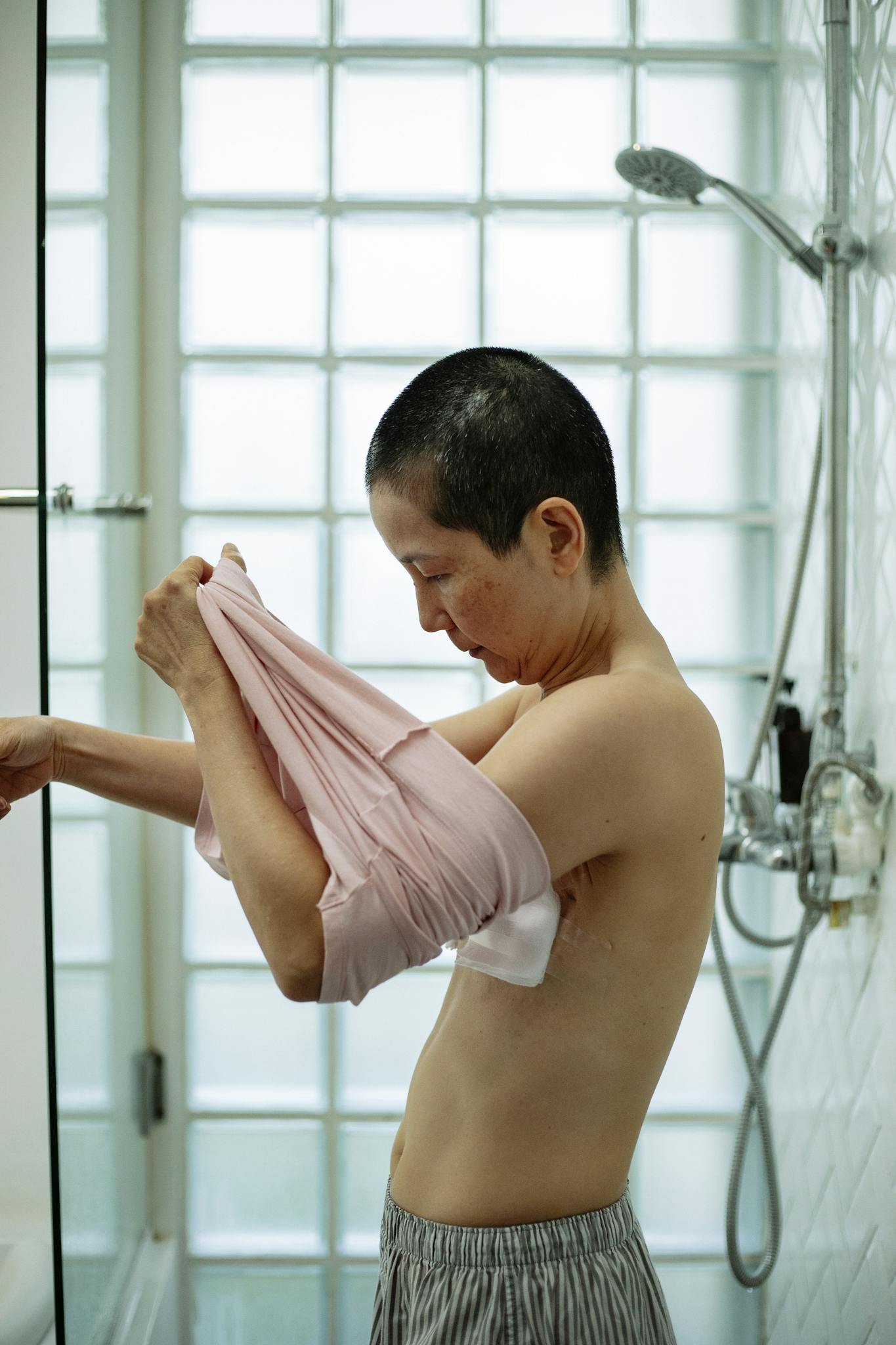 Side view of calm Asian female with short dark hair and sterile bandages on breast undressing in light bathroom